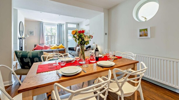 A dining area with a wooden table set for four, surrounded by white chairs. A vase with flowers is on the table, and there are wine glasses and a bottle. The room has white walls, a ceiling light, and a radiator. - 2 Bed in Whitstable (oc-f28381) (Whitstable)