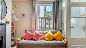 A cozy living room with a grey sofa adorned with pink and yellow cushions, a bookshelf, a small table with a lamp, and a window with curtains. - 2 Bed in Whitstable (oc-f28381) (Whitstable)