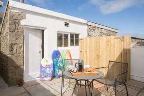 A small stone building with a white door, a window, and a wooden fence. There is a table with chairs and a surfboard leaning against the wall. - 3 Bed in Pendeen (oc-t22674) (Pendeen)