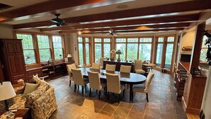A spacious living area with a dining table surrounded by chairs, a sofa, and a fireplace. Large windows provide natural light and views of the outdoors.