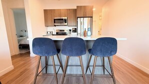 A modern kitchen with wooden cabinets, a microwave, and a refrigerator. There are three blue bar stools in front of the counter. - The Cache Contemporary DT Condo (Golden)
