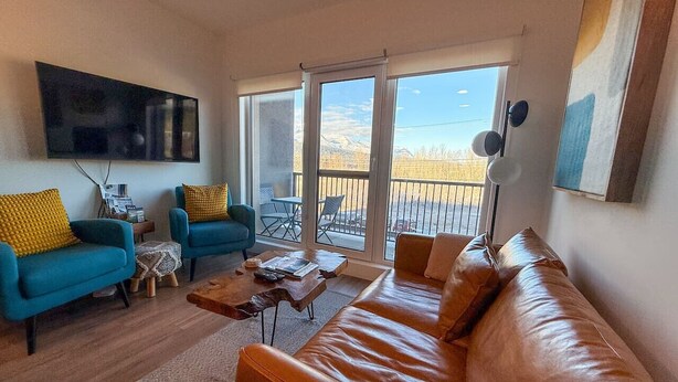 A living room with a brown leather sofa, a blue armchair, and a coffee table. There is a flat-screen TV mounted on the wall. - The Cache Contemporary DT Condo (Golden)