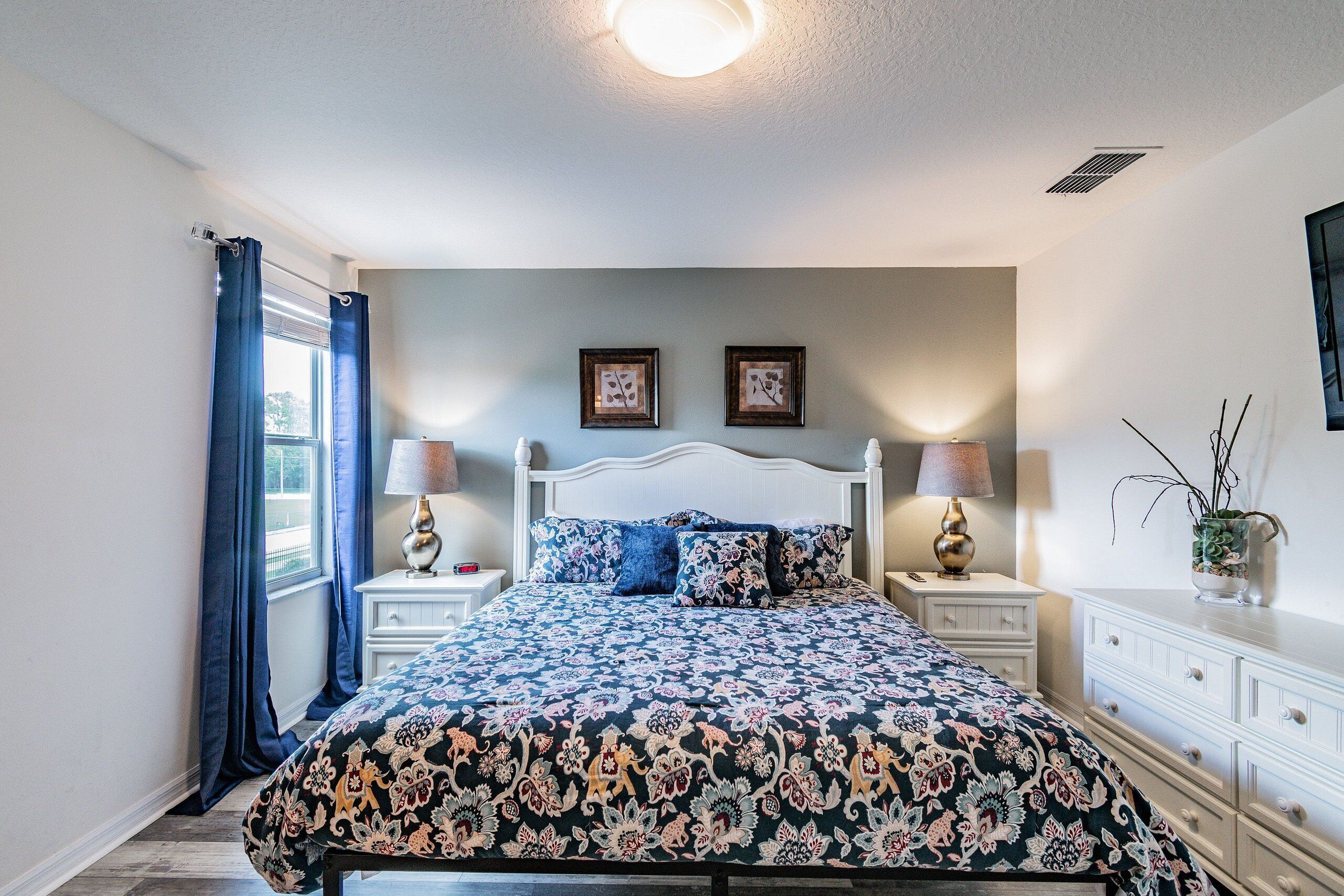 A bedroom with a large bed, two bedside tables with lamps, a dresser, and a window with curtains.