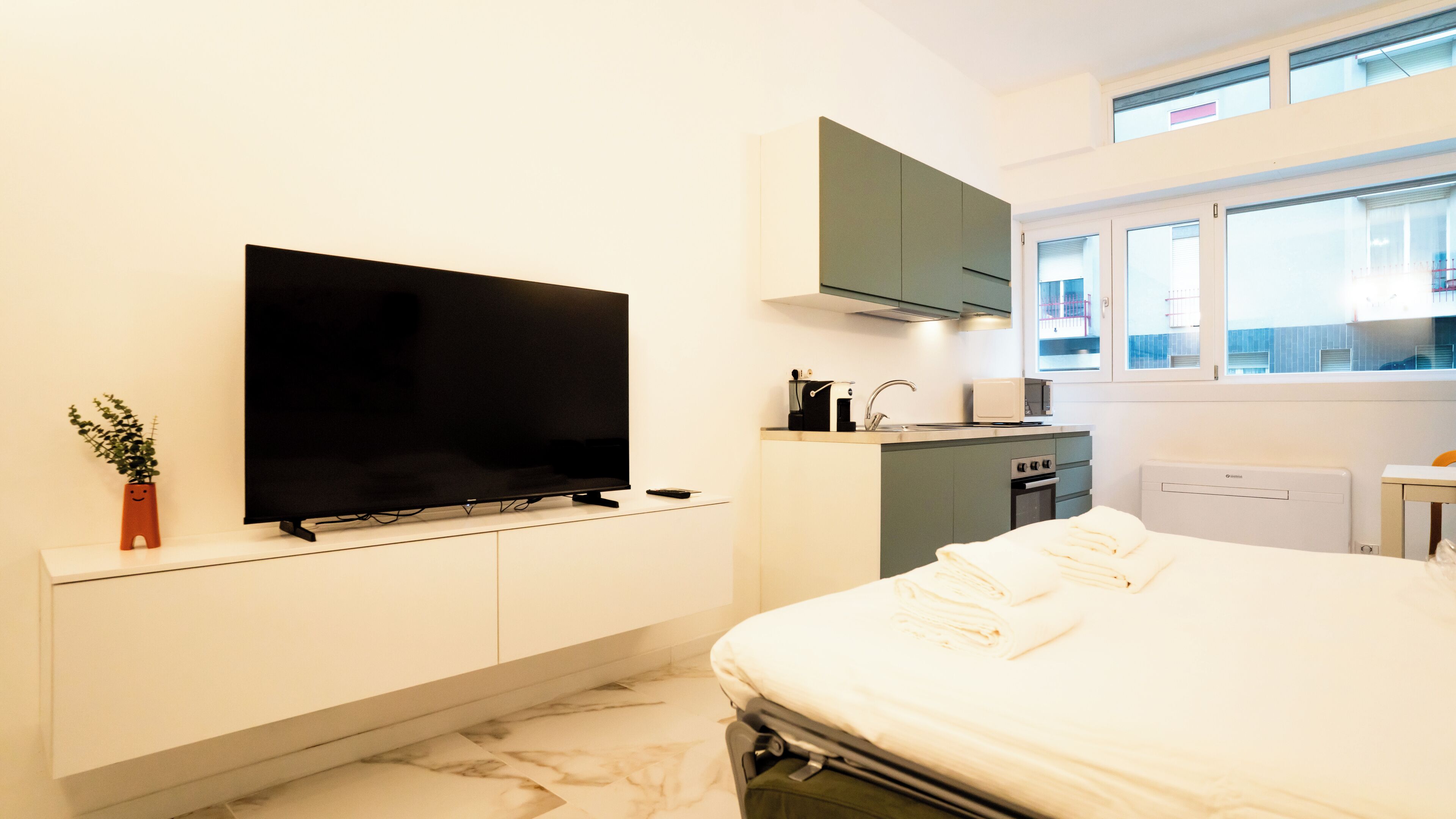 A compact hotel room with a bed, a television, a small kitchenette, and a window with a view of the outside.