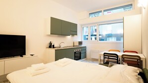 Hotellihuone, jossa on sänky, televisio, pieni keittiö ja ikkuna, josta on näkymä rakennuksiin.