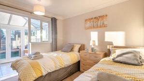 A bedroom with two beds, a window with curtains, a bedside table with a lamp, and a painting on the wall. - 2 Bed in Holt (oc-r28378) (Holt)