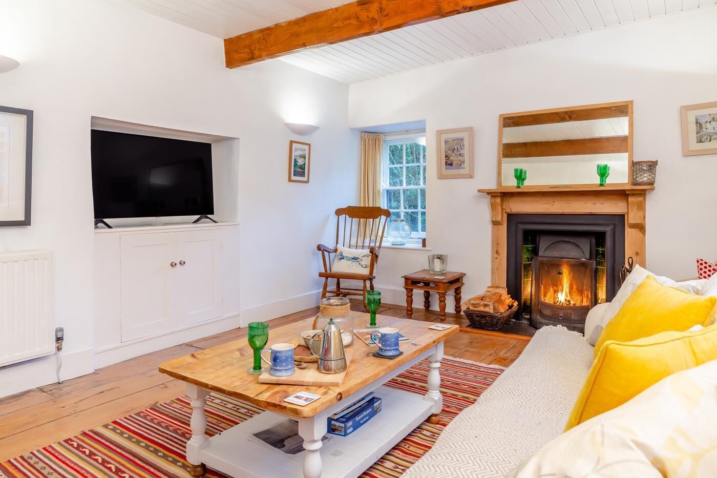 A cozy living room with a fireplace, a television, a wooden table with items on it, and a sofa with yellow cushions.