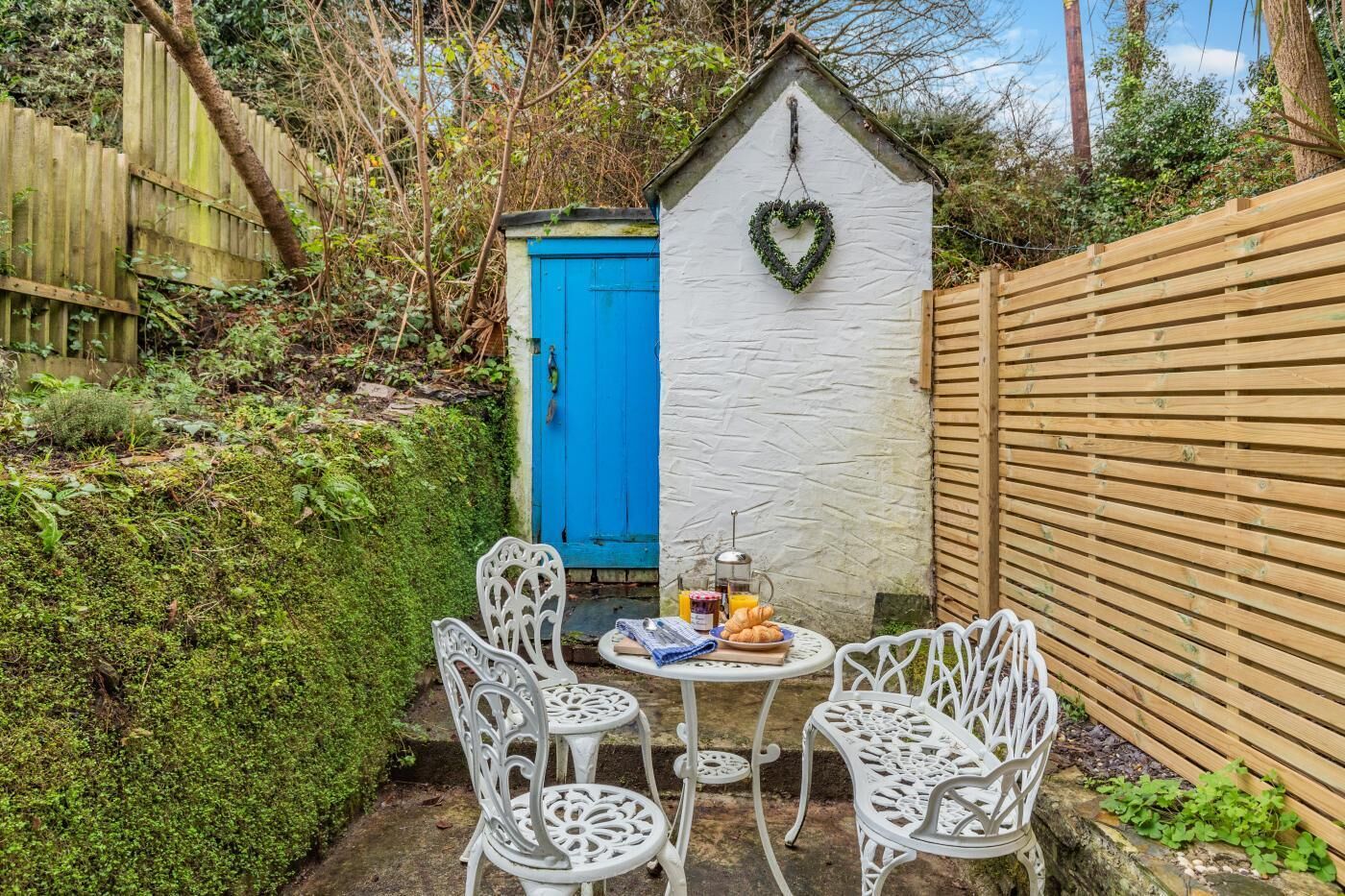 A small outdoor seating area with white metal chairs and a table, a blue door, and a heart-shaped decoration on a white wall.