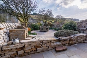 Ein von Grün und einer Steinmauer umgebener Außenbereich mit einer Terrasse, einem Tisch und Stühlen.