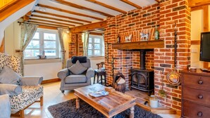 A cozy living room with exposed wooden beams, a brick fireplace, and a comfortable seating area with a sofa and armchair. - 3 Bed in Gressenhall (oc-s28418) (Gressenhall)