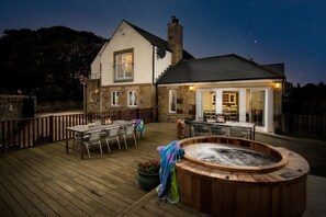 Eine Holzterrasse mit Whirlpool, Sitzgelegenheiten im Freien und einem Gebäude mit Naturstein-Fassade.
