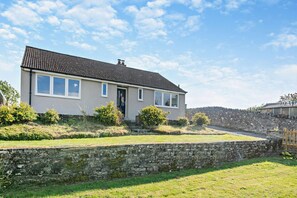 Une maison à deux étages, avec un toit en tuiles, un mur de pierre à l’avant et une pelouse verdoyante.