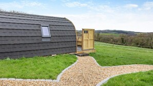A rustic cabin with a window and a wooden outhouse in a grassy field with a gravel path. - 1 Bed in Crediton (oc-p28229) (Crediton Hamlets)