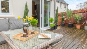 Une terrasse extérieure avec une table en verre, des meubles en osier, une cafetière, des tasses et des biscuits.