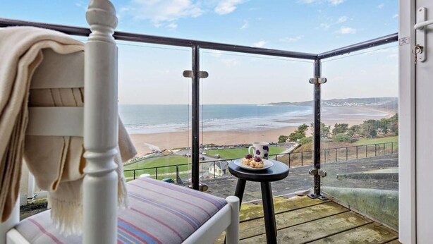 Een balkon met een bed, een klein tafeltje met een kopje en gebak, en uitzicht op het strand en de zee.