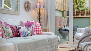 A cozy living room with a white sofa, floral cushions, a patterned rug, and a view of an outdoor patio with a grill. - 3 Bed in Padstow (oc-b28803) (Padstow)