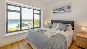 A bedroom with a large window, a bed with patterned bedding, a headboard, a bedside table with a lamp, and a painting on the wall. - 4 Bed in St. Ives (oc-c28788) (St Ives)