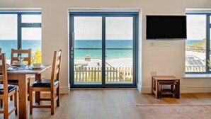 A room with a dining table set for two, a TV mounted on the wall, and a large sliding glass door leading to a balcony with a view of the ocean. - 4 Bed in St. Ives (oc-c28788) (St Ives)
