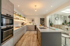 A modern kitchen with built-in appliances, a central island with a sink, and a dining area with a white table and chairs. - 5 Bed in Mitchell (oc-c28801) (Mitchell)
