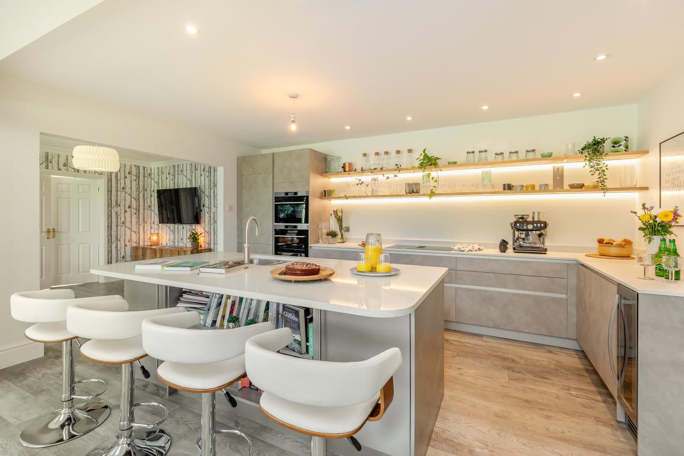 A modern kitchen with a central island, bar stools, a built-in oven, and a variety of kitchenware on the shelves.