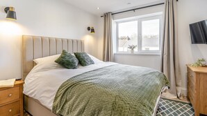 A bedroom with a bed, bedside lamps, a window with curtains, a TV on the wall, and a wooden cabinet. - 2 Bed in Mumbles (oc-c28839) (The Mumbles)