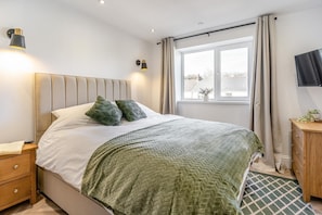 A bedroom with a bed, bedside lamps, a window with curtains, a TV on the wall, and a wooden cabinet. - 2 Bed in Mumbles (oc-c28839) (The Mumbles)