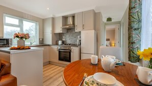 A modern kitchen with white cabinets, a central island, and a dining area with a wooden table set for tea. - 1 Bed in Beaworthy (oc-h28859) (Highampton)