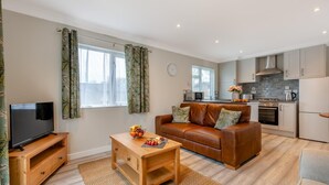 A modern living room with a flat-screen TV, a wooden coffee table with fruit, a brown leather sofa, and a kitchen with a stove, oven, and microwave. - 1 Bed in Beaworthy (oc-h28859) (Highampton)