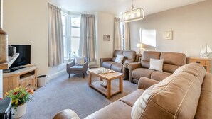A spacious living room with a large sofa, a smaller armchair, a coffee table, and a television on a wooden stand. - 4 Bed in Whitby (oc-b28403) (Whitby)