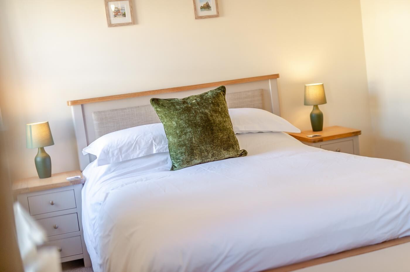 Un lit bien fait, avec du linge de lit blanc et un oreiller vert, flanqué de deux tables de chevet avec des lampes, dans une chambre simple avec une tête de lit en bois et des murs unis.