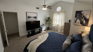 Ein Schlafzimmer mit einem großen Bett, einem Fernseher, einem Deckenventilator, einem Fenster mit Vorhängen und einem Bild an der Wand.