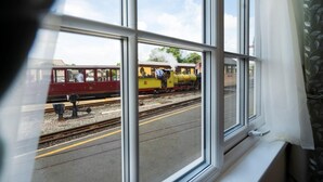 Veduta attraverso un finestrino di un treno sui binari, con un edificio e vegetazione sullo sfondo.