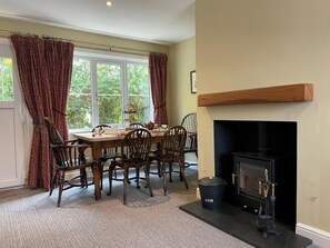 A dining area with a table set for four, surrounded by wooden chairs, a fireplace, and a window with curtains. - 2 Bed in Ravenglass (oc-h28842) (Ravenglass)