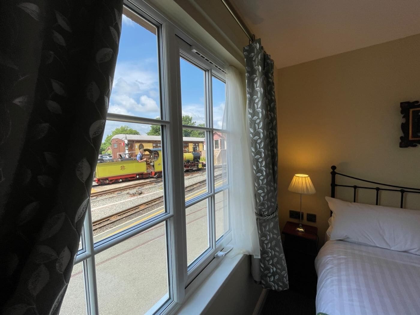 Ein Hotelzimmer mit einem Bett, einem Nachttisch, einer Lampe und einem Fenster mit Blick auf einen vorbeifahrenden Zug.