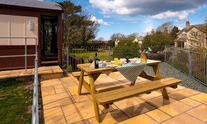 A picnic setup with a table and benches on a tiled patio, surrounded by a metal fence and a building. - 2 Bed in Ravenglass (oc-m28844) (Ravenglass)
