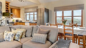 A modern living room with a sofa, dining table, and chairs. There is a kitchen area with wooden cabinets and a window with a view. - 3 Bed in Upottery (oc-t28828) (Upottery)