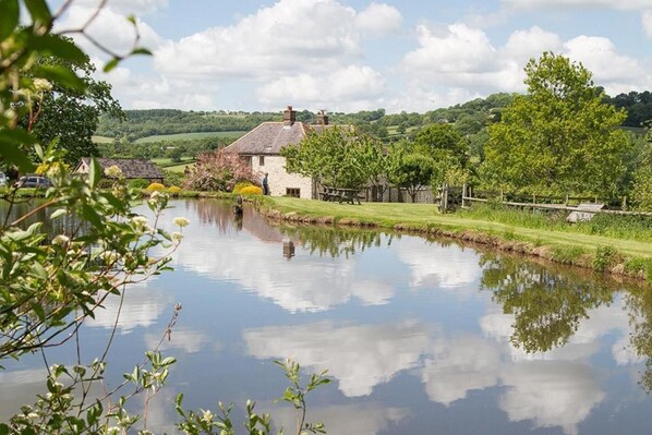 A serene riverside scene with a stone house, lush greenery, and a clear sky. - 3 Bed in Upottery (oc-t28827) (Upottery)