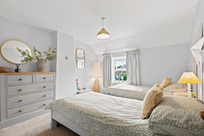 Ein Schlafzimmer mit zwei Betten, einem Schminktisch, einem Spiegel, einer Lampe und einem Fenster mit Vorhängen.