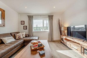 Un salon moderne avec un canapé en cuir, une table basse en bois sur laquelle sont disposés des en-cas, un téléviseur à écran plat et une grande fenêtre agrémentée de rideaux.