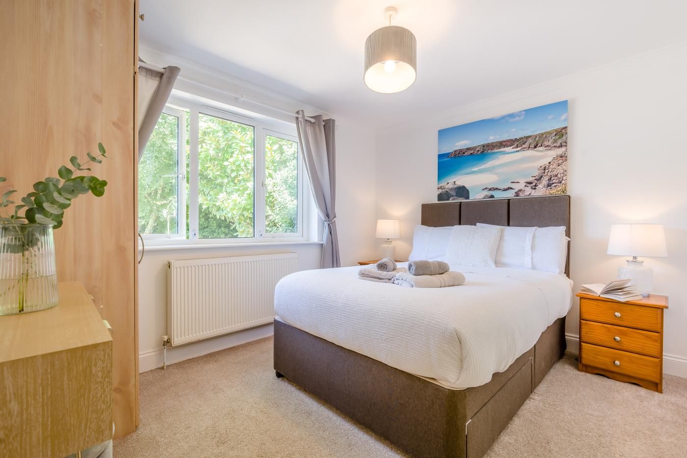 A bedroom with a bed, bedside tables, a window with curtains, a radiator, and a painting on the wall.