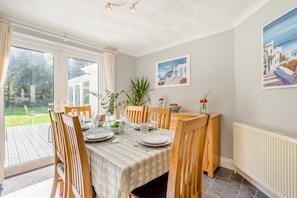 Ein Essbereich mit Tisch für vier Personen, Holzstühle, Blick auf den Garten durch große Fenster und ein Bild an der Wand.