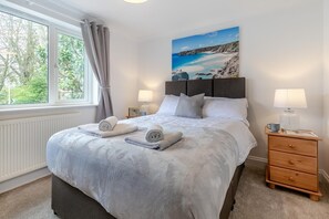 Une chambre à coucher avec un grand lit, deux tables de chevet avec des lampes, une fenêtre avec des rideaux et un tableau représentant une scène de plage.