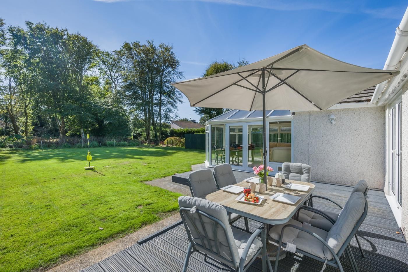 An outdoor patio with a table and chairs, a large umbrella, and a view of a garden and house.