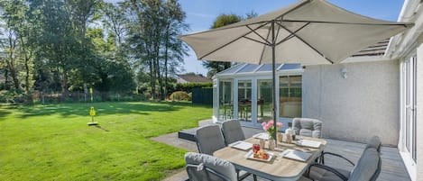 An outdoor patio with a table and chairs, a large umbrella, and a view of a garden and house.