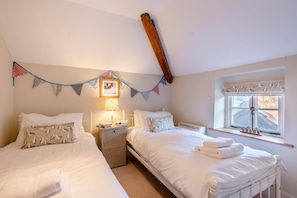 A room with two beds, a bedside table with a lamp, a window with curtains, and a wall with bunting. - 2 Bed in Langham (oc-t28836) (Langham)