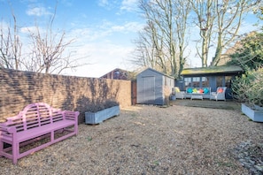 Ein Garten mit einer rosa Bank, einem Gerätehaus und einer Sitzgruppe mit bunten Kissen.