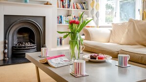Un salon chaleureux avec une cheminée, une bibliothèque, une table basse avec des livres et un vase de fleurs, ainsi qu’un canapé.