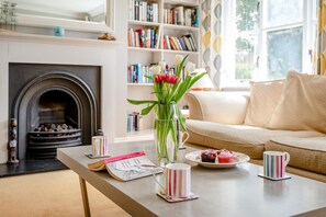 Ein gemütliches Wohnzimmer mit Kamin, Bücherregal, Couchtisch mit Büchern und einer Blumenvase sowie einer Couch.