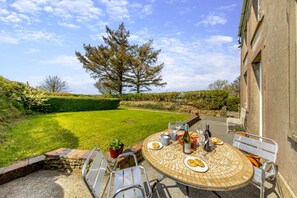 A patio with a round table set for dining, surrounded by chairs, with a view of a green lawn and a large tree in the background. - 3 Bed in Llanrhystud (oc-m28913) (Llanrhystyd)