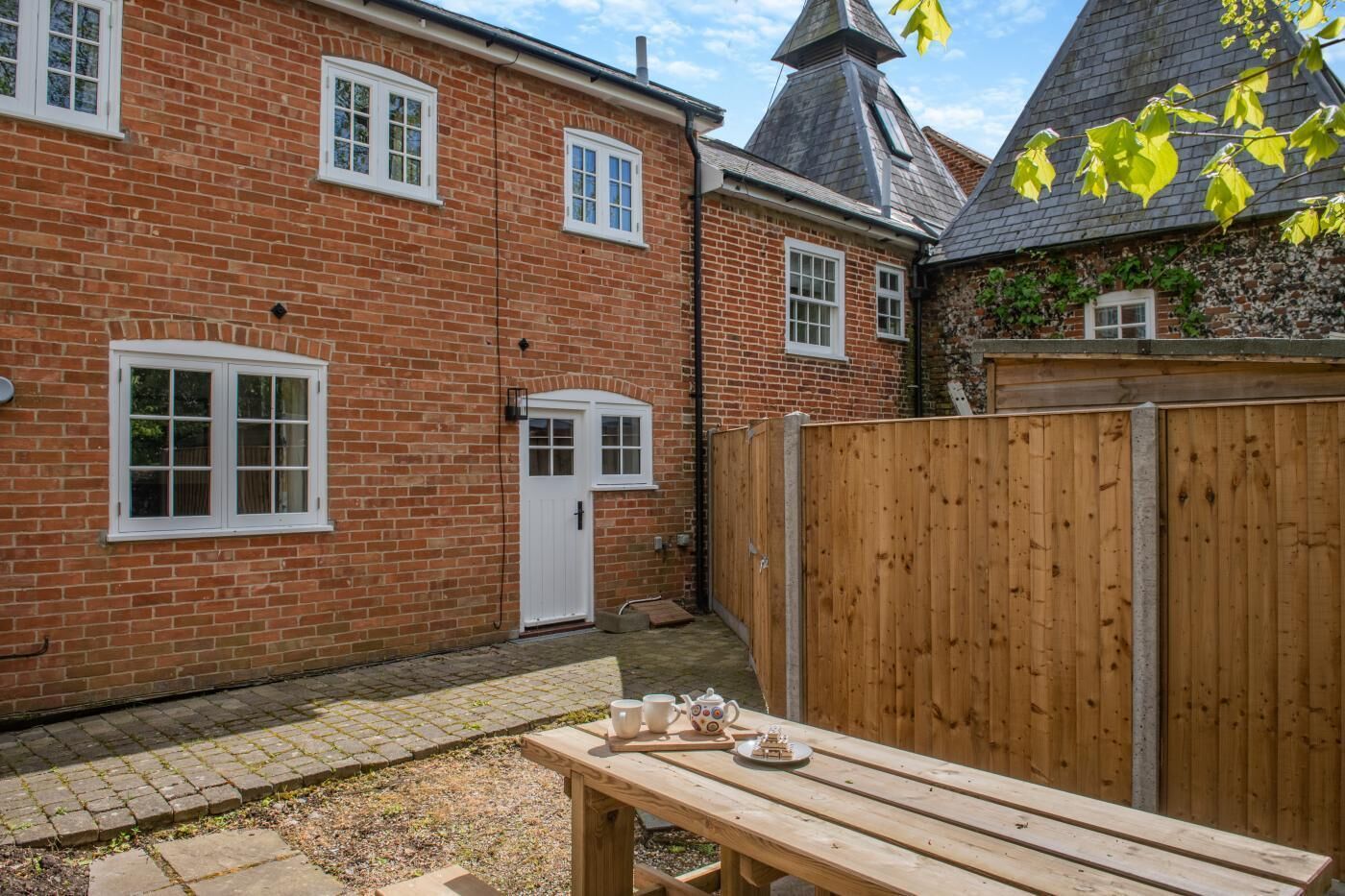 A brick house with a wooden fence, a door, and a picnic table with cups and a teapot.